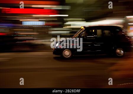 Ein schwarzes Londoner Taxi in der Nacht mit Bewegungsunschärfe gegen verschwommene Neonlichter Stockfoto