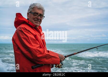 Porträt eines alten Mannes, der Freizeit genießt und auf den Felsen am Meer angeln kann. Angelkonzept. Stockfoto