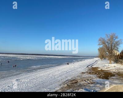 Blick von der Böschung auf die gefrorene Wolga in Samara, Russland. In der Ferne laufen die Menschen an einem sonnigen Tag auf dem Eis Stockfoto