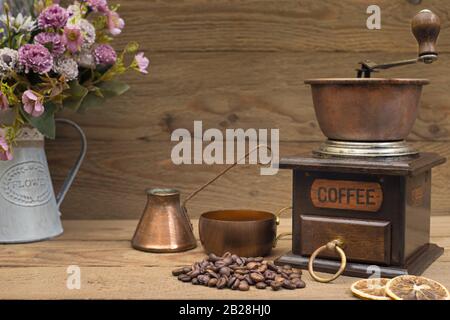 Vintage manuelle Kaffeemühle mit Kaffeekanne, Kaffeebohnen, retro Kupfer Kaffeetasse auf einem hölzernen Hintergrund und Vase mit Blumen. Stockfoto