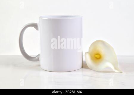 Leerer Becher mit weißer Tasse, 11 oz. Weißer Kaffeetasse neben einer schönen weißen Calla-Lilie aus Ostern. Stockfoto