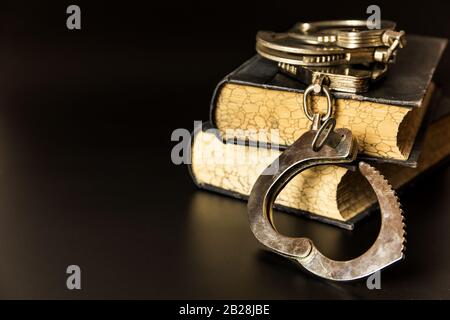 Polizeihandschellen und Gesetzbücher auf schwarzem Hintergrund. Holzgavel und Bücher auf Holztisch, Gesetzeskonzept. Recht und Ordnung. Stockfoto