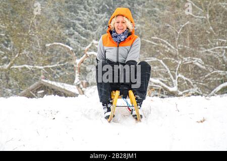 Blonde Frau mit Locken und orangefarbener Jacke auf einem Schlitten im Schnee Stockfoto