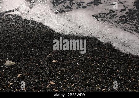 Flacher vulkanisch schwarzer Strand und Kiesel, Wasserwellen und weißer Schaum wäscht die Küste mit nassem dunklem Sand an der Meeresküste. Ocean Shore Natur Stockfoto