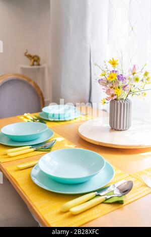 Buntes Set mit Geschirr und einem Blumenstrauß aus wilden Blumen auf einem braunen Holz-Esstisch in natürlichem Licht, angewinkelt. Stockfoto