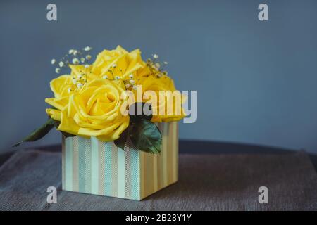 Gelber Rosenstrauß mit grünen Blättern im Kasten auf grauem Grund. Geschenkidee für Frauen oder Muttertage. Festlicher Hintergrund mit Kopierbereich Stockfoto