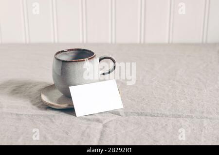 Tasse Kaffee und leeres Geschäft, Platzkarte. Moody Frühstück Tisch Schreibwaren Spockup Szene. Beige Leinen Tischdecke Hintergrund. Spärliches Stillleben Stockfoto