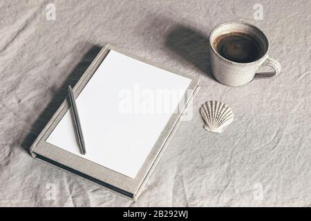 Tasse Kaffee, Buch, leeres Blatt Papier und silberner Stift. Moody Frühstück Tisch Schreibwaren Spockup Szene. Beige Leinen Tischdecke Hintergrund. Spärlich Stockfoto