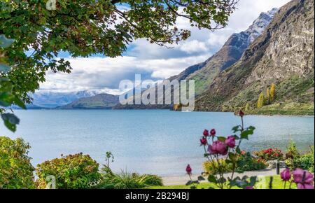 Blick durch einen Rahmen von Bäumen und Rosen auf den Lake Wakatipu und schneebedeckte Alpenberge im Herbst in Neuseeland Stockfoto