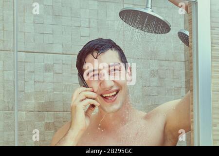 Der lachende weiße Mann benutzt ein Smartphone, das unter den Wasserläufen steht, die aus der Dusche strömen. Ein junges kaukasisches Männchen spricht am Telefon im B. Stockfoto