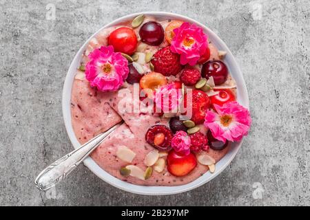 Schüssel aus rosafarbenem Beerensmoothie oder schöner Sahne aus gefrorenen Früchten und Nüssen mit frischen Beeren, Nüssen und Rosenblumen. Draufsicht. Gesunder romantischer Breakfa Stockfoto