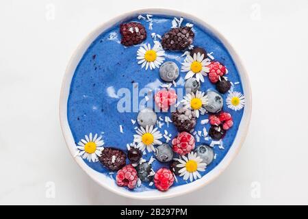 Blaue Spirulina-Smoothie-Schüssel mit frischen und gefrorenen Beeren, Kokosnuss- und Kamillenblüten. Gesundes Frühstücksdessert. Draufsicht Stockfoto