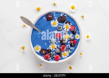 Gesundes Frühstücksdessert. Blaue Spirulina-Smoothie-Schüssel mit frischen und gefrorenen Beeren, Kokos- und Kamillenblüten mit Löffel auf weißem Marmortisch. Stockfoto