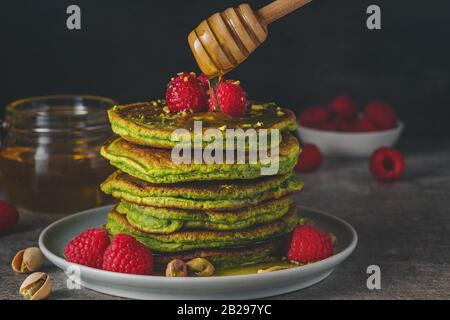 Matcha-Tee-grüne Pfannkuchen. Haufen hausgemachter Pfannkuchen mit frischen Himbeeren, Pistazien und fließendem Honig auf dunklem Hintergrund. Niedriger Schlüssel. Gesundes Frühstück Stockfoto