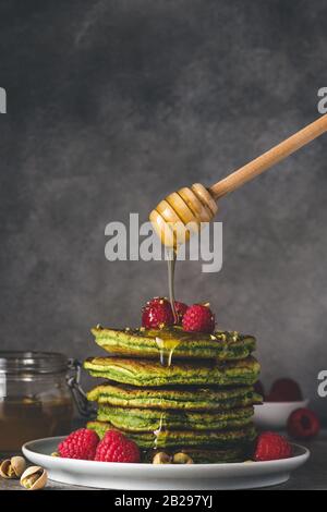 Hausgemachte Matcha-Pfannkuchen mit frischen Himbeeren, Pistazien und fließendem Honig auf dunklem Hintergrund. Niedriger Schlüssel. Gesundes Frühstücksdessert. Vertikale Orienta Stockfoto