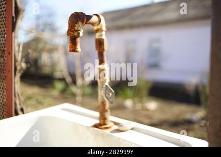 Altes rostiges Wasser trank auf einem Waschbecken in einem ländlichen Hof. Die Fassade des Hauses im Hintergrund verwackelt. Sonniger Tag Stockfoto