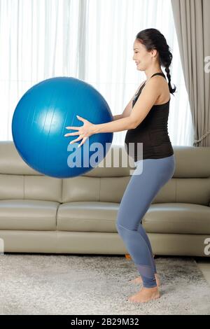Fröhliche junge schwangere Vietnamesin, die mit Yoga-Ball in den Händen hockt Stockfoto