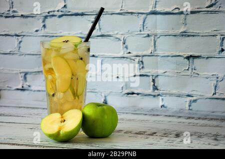 Eine große Glasschale mit Saft, Eis und Fruchtscheiben, neben einem ganzen und von einer Ziegelwand auf einem Holztisch grünen apfel schneiden. Nahaufnahme. Stockfoto