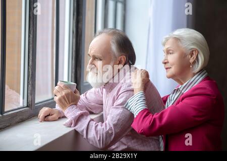 Älteres grauhaarige Paar, das in der Nähe des Fensters steht und nachdenklich aussieht Stockfoto