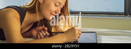 Junge Frau zieht vor dem Hintergrund eines Fensters mit Meerblick auf eine Tablette. Designer, Banner skizzieren, LANGFORMAT Stockfoto