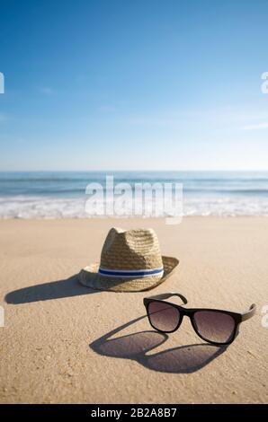 Einfacher Sommerhut und kühle Sonnenbrille, die lange Schatten in der untergehenden Sonne auf dem glatten Sand eines leeren Strandes werfen Stockfoto