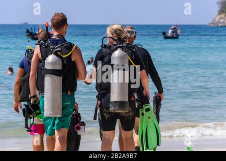 Vier Taucher mit Sporttanks gehen in Richtung Meer am Kata Beach, Phuket, Thailand Stockfoto