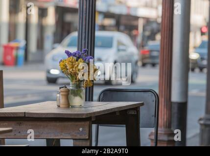 Ein bunter kleiner Blumenstrauß sitzt in einem Krug auf einem rustikalen Holztisch vor einem Café in der belebten Brunswick Street in Melbourne, Australien Stockfoto