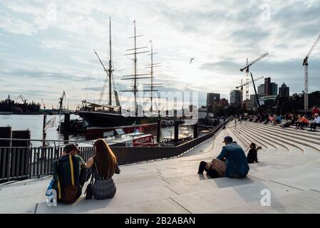 Hamburg - 4. August 2019: Menschen, die im Sommer auf der Promenade sitzen und den Sonnenuntergang im Hamburger Hafen beobachten Stockfoto