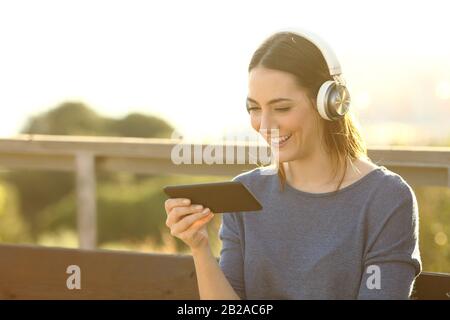 Fröhliche Frau, die bei Sonnenuntergang Videos mit Headset auf ihrem Smartphone im Park sieht Stockfoto