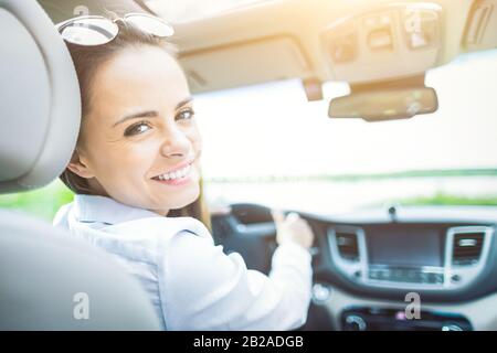 Schöne junge Frau, die mit einem Lächeln im Innenraum eines neuen Autos sitzt. Er ist froh, dass er ein Fahrer ist. Stockfoto