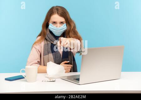 Mädchen in hygienischer Maske sitzt am Schreibtisch mit Laptop und zeigt auf Kamera, Warnung, die Ratschläge gibt, Schutzfilter gegen ansteckende Krankheit zu tragen, Stockfoto