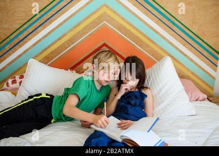 Der Junge sitzt auf einem Bett und hilft seiner Schwester bei ihren Hausaufgaben Stockfoto