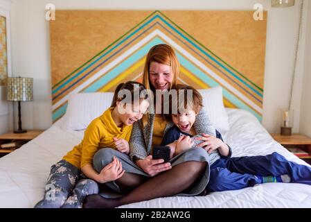 Fröhliche Mutter sitzt mit zwei Kindern auf dem Bett und schaut auf ein Handy Stockfoto