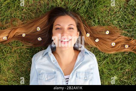 Nahaufnahme des hübschen Mädchens, das auf Gras mit Gänseblümchen in ihren Haaren liegt, blickt im Frühjahr auf die Kamera Stockfoto