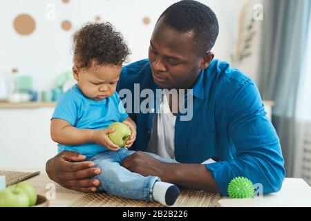 Porträt eines glücklichen afroamerikanischen Vaters, der mit niedlichem kleinen Jungen auf dem Tisch mit grünen Äpfeln im Innenbereich spielt, Kopierraum Stockfoto