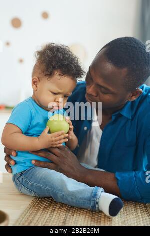Porträt eines glücklichen afroamerikanischen Vaters, der mit niedlichem kleinen Jungen auf dem Tisch mit grünen Äpfeln im Innenbereich spielt Stockfoto