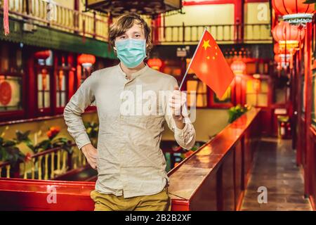 Urlaub in China genießen. Junger Mann mit chinesischer Flagge in medizinischer Maske auf chinesischem Hintergrund. Reise nach China Konzept. Touristen fürchten 2019 Stockfoto