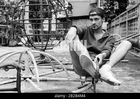 Junger bärtiger Mann denken, während an der alten rostigen Merry go round sitzen und das Tragen von Gap in den alten Spielplatz Stockfoto