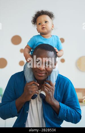 Vertikales Porträt eines glücklichen afroamerikanischen Mannes, der Sohn auf den Schultern trägt und lächelnd mit der Kamera blickt Stockfoto