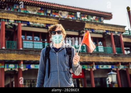 Urlaub in China genießen. Junger Mann in medizinischer Maske mit nationalchinesischer Flagge auf dem Hintergrund der alten chinesischen Straße. Reise nach China Konzept Stockfoto