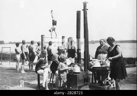 Camper versammeln sich um eine Kochzone mit mehreren Öfen und bereiten im Zeltlager Tegel am Tegel Essen zu. Inzwischen beobachten die Männer, wie einer von ihnen auf einem horizontalen Balken ausübt. Stockfoto