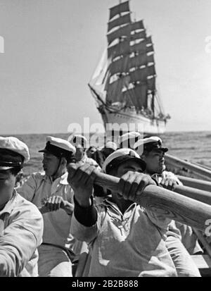 Kadetten der japanischen Marine sitzen während einer Übung in einem Rettungsboot auf offener See und rudern. Ihr Ausbildungsschiff Taisei Maru ist im Hintergrund verankert. Stockfoto