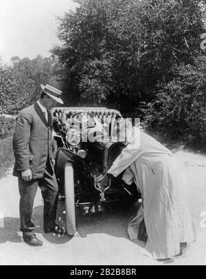 Eine Frau, die einen Smock trägt, um nicht schmutzig zu werden, startet den Motor ihres Autos mit einer Handkurbel, während der Mann zu ihrer linken sie beobachtet. Stockfoto