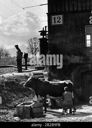 Wegweiser in der Nähe eines Stationsgebäudes auf dem Land. Davor melkende Frau eine Kuh. Stockfoto