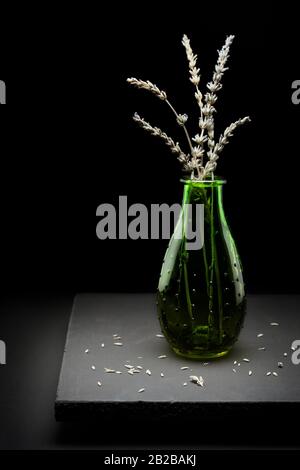 Getrockneter Lavendel in einer grünen Flasche, die im Studio fotografiert wurde, mit hellem, dunklem Licht. Blumen fielen auf die Plattform, um Realismus zu erzeugen Stockfoto