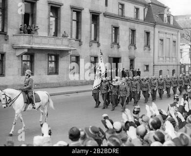 Das österreichische Bataillon marschiert an der neuen Staatskanzlei vorbei. Adolf Hitler steht auf dem Balkon und macht den Nazi-Salut. Der führende Soldat trägt die Flagge der Kaiserlichen Kaiserlichen Kaiserlichen Trabantgarden. Sie werden von Passanten am Straßenrand mit dem Nazi-Salut begrüßt. SS-Männer stehen an einem Seiteneingang der Staatskanzlei. Stockfoto