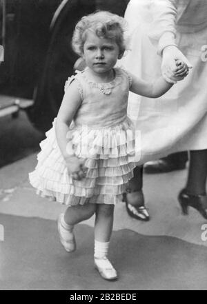 Elizabeth II. Spaziert als Kleinkind, während sie die Hand eines Erwachsenen hält. Sie trägt ein Rüschenkleid und eine Perlenkette. Stockfoto