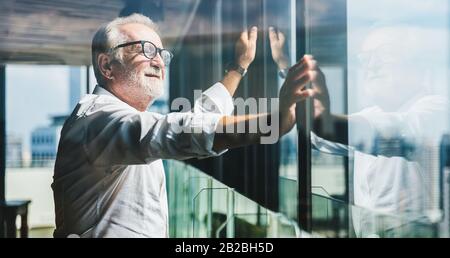 Ruhestandskonzept. Ein hochrangiger, grauhaariger Geschäftsmann, der in der modernen Business Lounge hoch oben in einem Büroturm steht und nach rechts schaut. Stockfoto