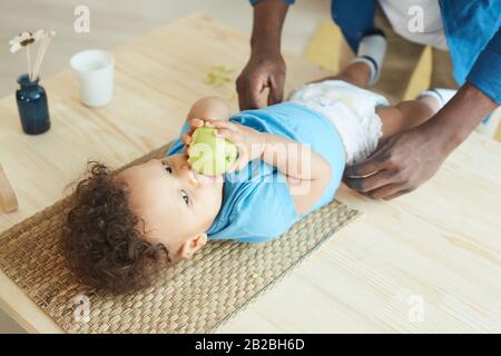 Hochwinkelige Ansicht bei nicht erkennbarem Vater, der Windel in süße afroamerikanische Kleinkinder wechselt, die apple essen, Kopierraum Stockfoto