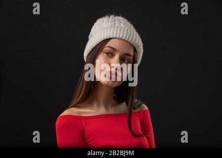 Das Porträt der schönen jungen europäischen Frau hält Lippen rund, trägt einen warmen Winterhut, stellt sich über schwarzem Hintergrund dar. Kaltwetterkonzept Stockfoto